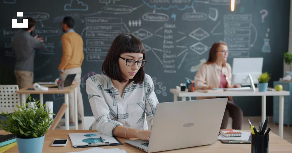 Young woman working on a laptop at a desk with a plant, while colleagues collaborate in the background against a chalkboard filled with diagrams and notes, illustrating a creative web design and development environment.