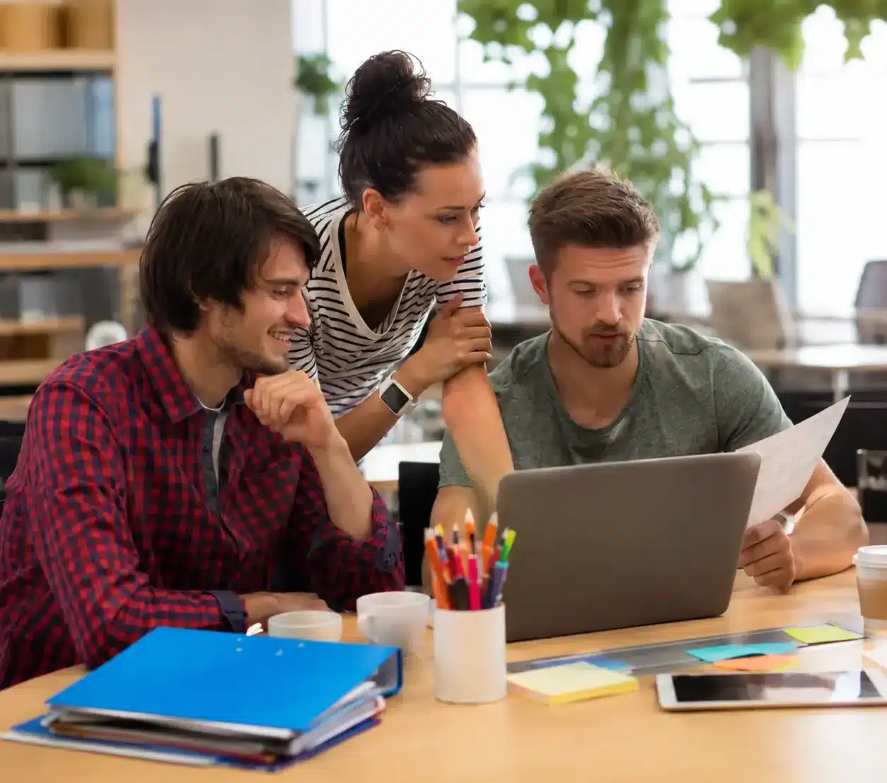 Three web design professionals collaborating around a laptop in a bright modern office, reviewing website layouts, project documents, and digital ideas. The contemporary workspace features natural light, notebooks, sticky notes, and a collaborative environment focused on web development, UX design, and creative problem-solving.